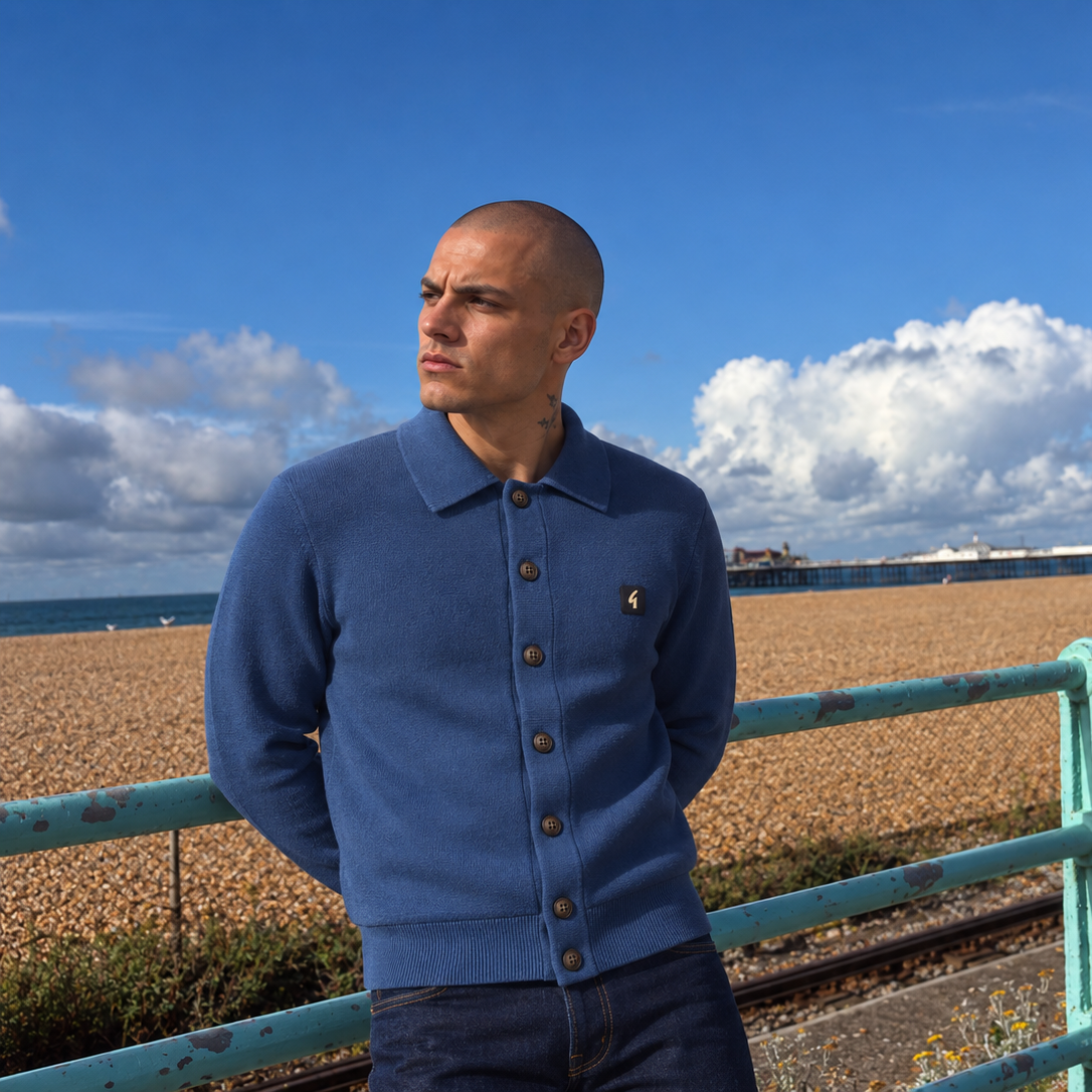 A man wearing a blue knit polo-button cardigan sweater and dark jeans stands outdoors near a turquoise railing with a pebble beach and blue sky in the background.
