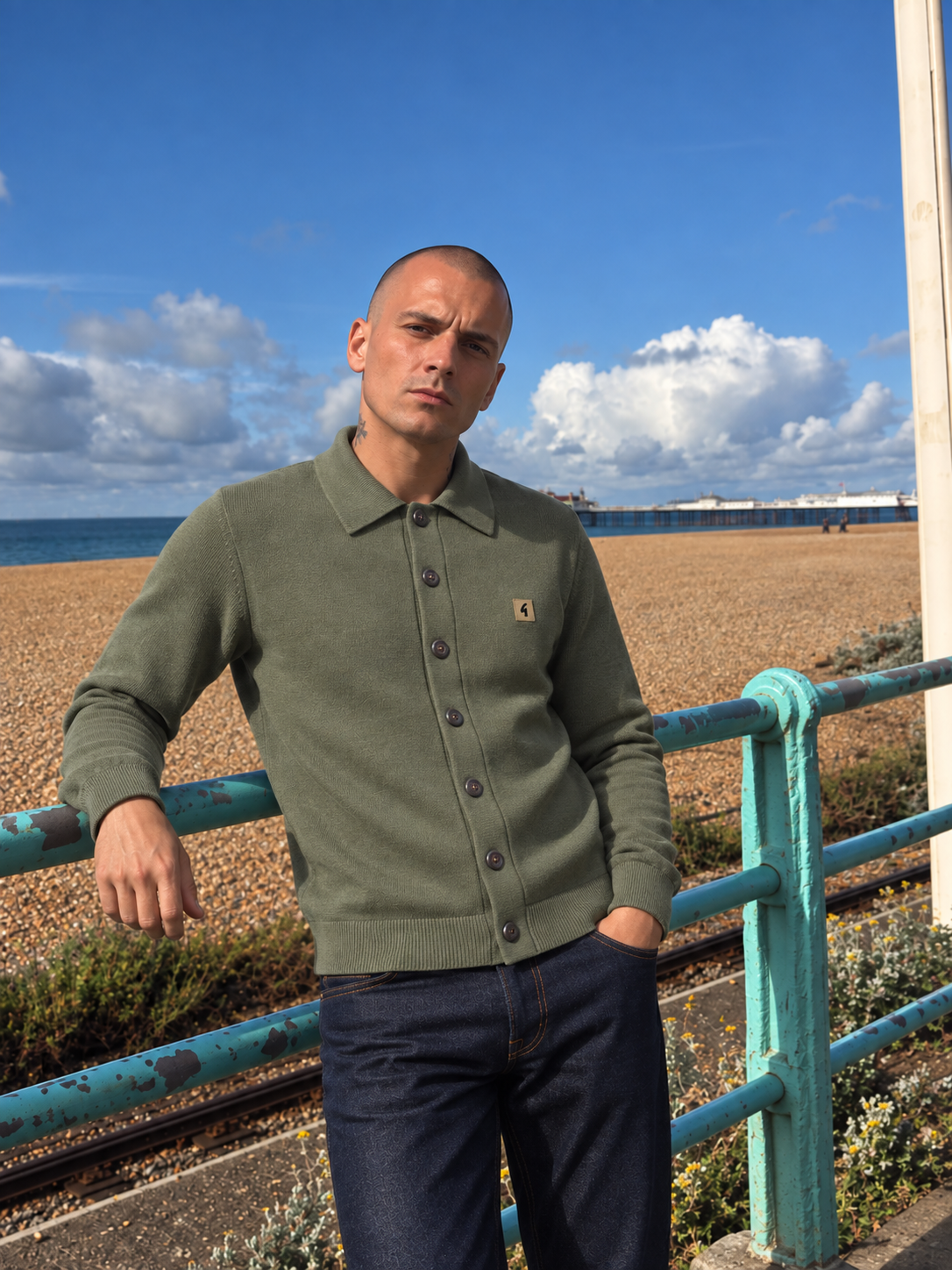 A man wearing an olive green knit polo cardigan with dark buttons and a small Carhartt logo patch, standing outdoors by a teal metal railing with a beach and blue sky in the background.