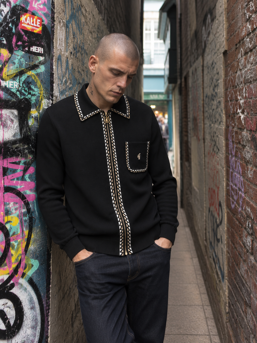A man wearing a black knit zip-up sweater with gold chain-stitched trim and a chest pocket stands in a narrow urban alleyway with graffiti-covered walls.