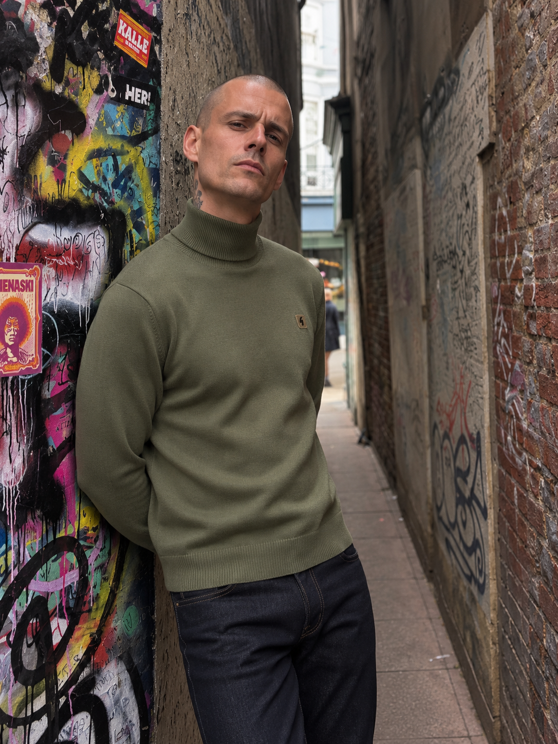 A man wearing an olive green knit turtleneck sweater and dark jeans leans against a graffiti-covered wall in a narrow urban alleyway.
