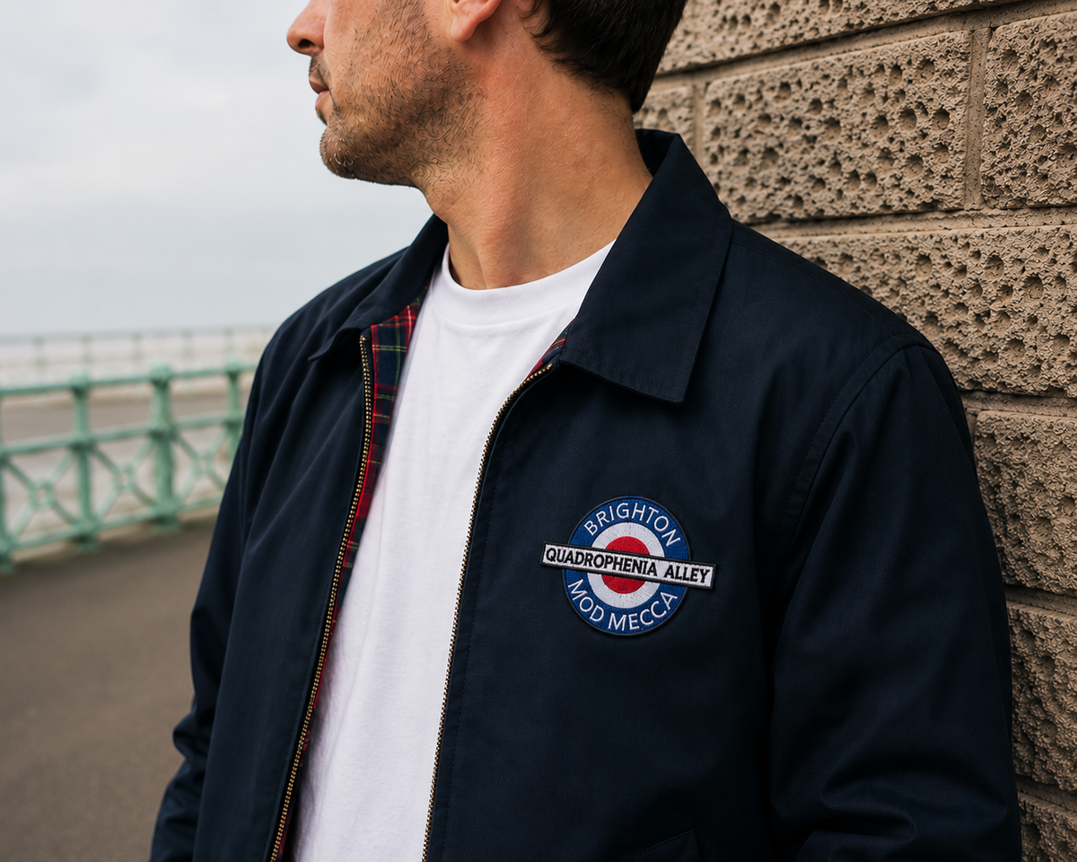 A man wearing a navy blue bomber jacket with a circular Brighton Quatrophaena Alley 400 Mecca patch on the chest, standing against a textured stone wall.