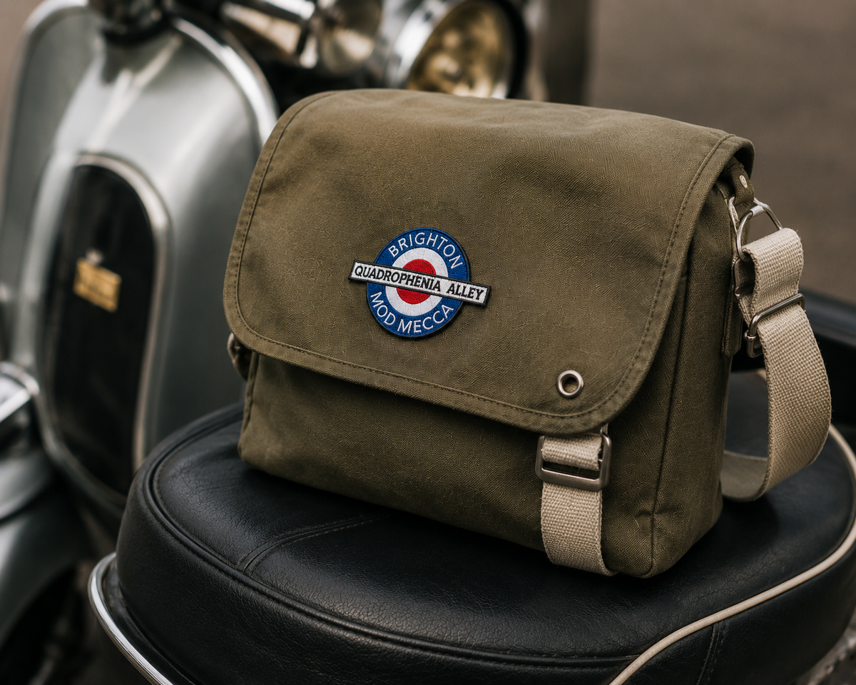 An olive green canvas messenger bag with a beige strap, featuring a round 'Brighton and Hove Motor Cycle Club' patch on the front flap, resting on a black leather motorcycle seat.